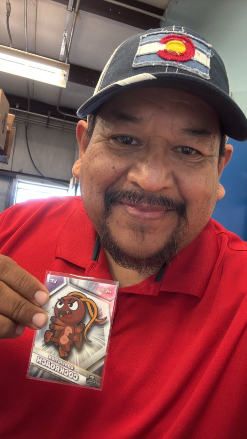 A man wearing a red shirt and a baseball cap with a Colorado flag patch smiles while holding up a packaged 'Cockroach Companion' trading card featuring a cartoon bug.