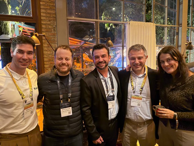 A photo of five people posing together at what appears to be a conference or networking event, all wearing name badges and lanyards, taken at night with warm lighting.
