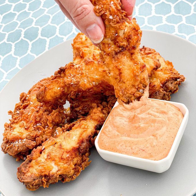 A hand dips a golden, crispy fried chicken tender into a small square bowl of creamy orange sauce, served on a light gray plate set against a blue patterned tablecloth—perfectly illustrating the simple pleasure of a "nice dip."