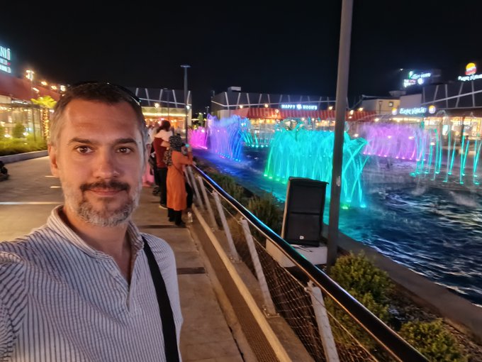 A nighttime selfie of a bearded man in a striped shirt standing in front of an illuminated musical fountain display with colorful lights and restaurants in the background.