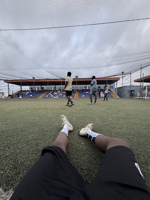 A first-person perspective shot looking down at a person's legs and cleats resting on a grass field, with other people playing sports in the background under a large net.