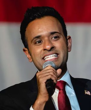 A close-up photograph of Vivek Ramaswamy smiling while speaking into a microphone, wearing a dark suit, light blue shirt, and red tie with an American flag pin.