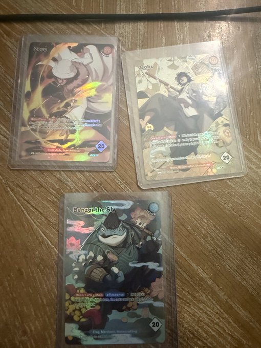 Three holographic Azuki TCG leader cards—Zoro, Babu, and Benzaiten the Sly—each showing a "20" value, displayed on a wooden table as featured pulls from the collection.