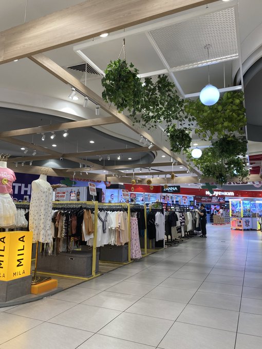 An indoor shopping mall scene with a clothing kiosk featuring apparel on yellow racks, overhead hanging plants, and a person walking in the distance near store signs for 'JANNA' and 'eyesum'.