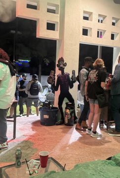A casual indoor snapshot showing a group of people gathered in what appears to be a lobby or hallway, with one person wearing a dark costume or mask and a large black bag on the floor.