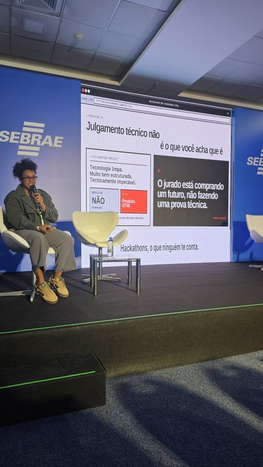 A woman speaks into a microphone on stage at a SEBRAE event, with a large screen behind her displaying a presentation titled "Julgamento técnico não é o que você acha que é" and "Hackathons, o que ninguém te conta," referencing technical judgment and hackathons during a Blockchain Rio gathering.