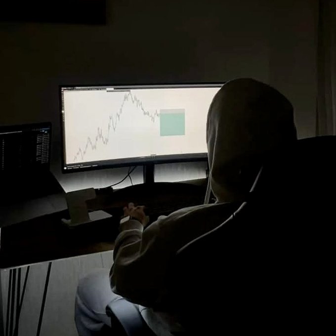 A silhouette of a person in a hoodie sitting in a dark room, looking at a bright computer monitor displaying a stock trading chart.