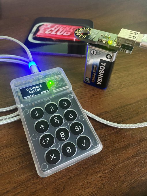 A Coinkite Coldcard Mk4 hardware wallet, powered by a Toshiba 9V battery and connected via USB, sits on a wooden surface displaying "Coldcard Wallet" on its screen. The image visually represents a Bitcoin hardware security device, referencing the accompanying post's unsubstantiated claims about potential brute-force attacks draining funds from Coldcard users.