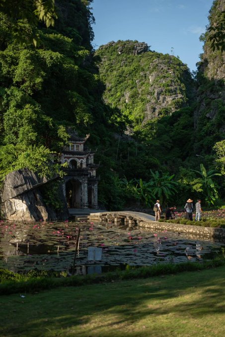 A scenic landscape featuring a traditional Asian stone gateway nestled between large, lush green karst mountains, with a lotus pond in the foreground where a few people are standing.