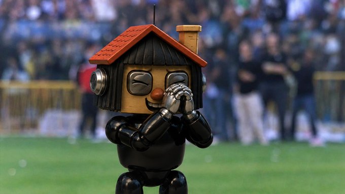 A small robot with a vintage house-shaped head featuring a red roof, black hair-like panels, and a painted smile stands on a green sports field with its metallic hands clasped together, with a blurred crowd in the background.