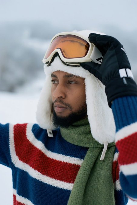 A close-up photograph of a man wearing a white furry winter hat, orange ski goggles, a striped sweater, and black gloves, standing in a snowy landscape.
