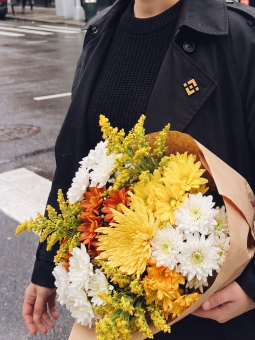 A person in a black trench coat holds a vibrant bouquet of yellow, white, and orange chrysanthemums wrapped in brown paper, with a small gold "21" pin visible on the coat lapel—evoking the arrival of spring in a city street setting.