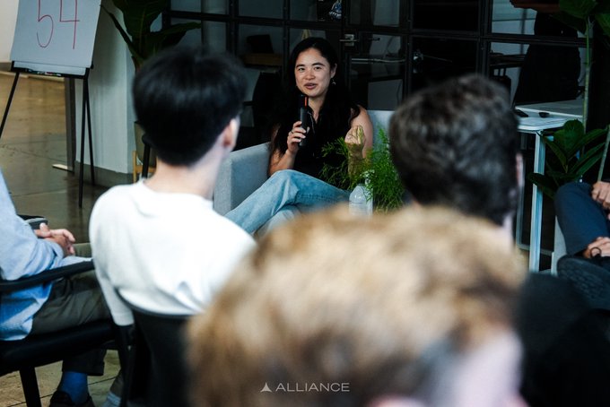 A woman speaks into a microphone during a panel discussion, seated while an audience listens, with the 'ALLIANCE' watermark visible at the bottom.