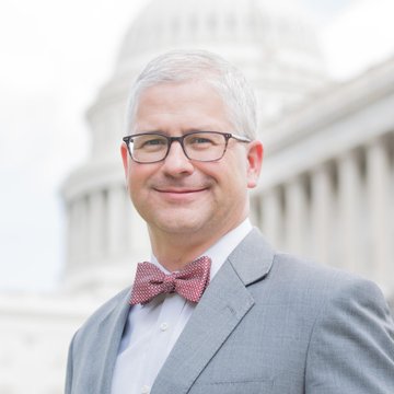 A man with glasses and a red bow tie smiles in front of the U.S. Capitol building. The image accompanies news about Patrick McHenry discussing the CLARITY Act's significance in technology legislation.