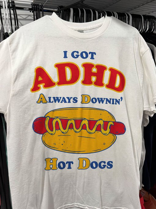 A white t-shirt hanging on a rack featuring a cartoon hot dog graphic and the text 'I GOT ADHD ALWAYS DOWNIN' HOT DOGS'.