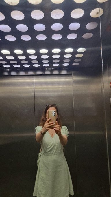 A woman in a light green dress takes a mirror selfie in an elevator with a perforated metallic ceiling.