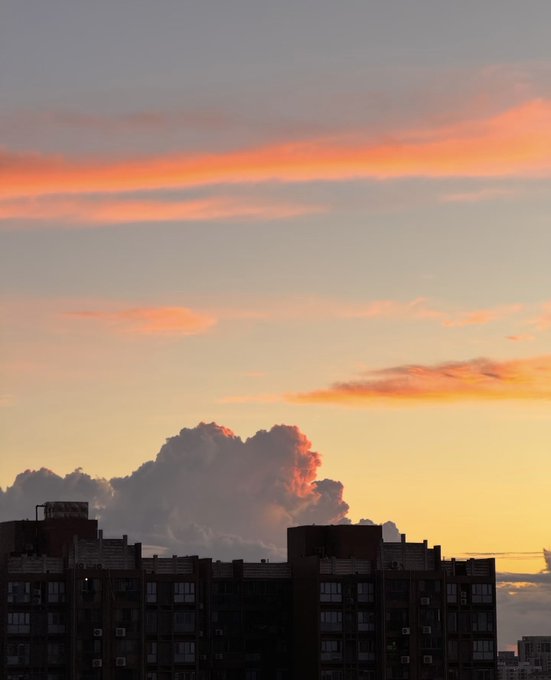 A dramatic sunset sky with pink and orange clouds towers above the silhouette of city buildings, with a large cumulus cloud glowing in the warm light.