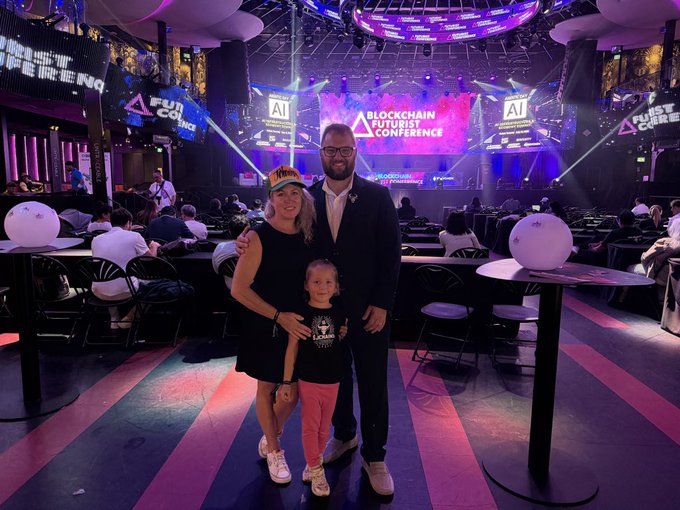 A family poses together at the "Blockchain Futurist Conference," with vibrant stage lighting and "AI" and "FUTURIST CONF" text visible in the background, capturing a memorable moment at the tech event that the caption describes as nicely summed up.