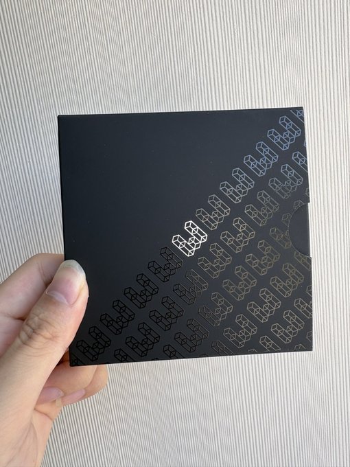 A hand holding a black square box with a repeating pattern and a small logo in the center, held against a textured white background.