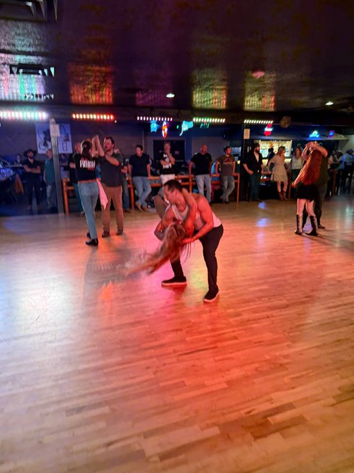 Two dancers perform a dip on a wooden floor in a dance hall, with other couples dancing and onlookers visible in the background under colorful lights.