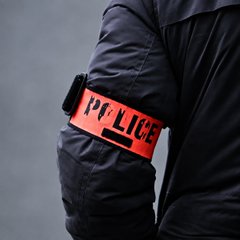 A close-up of a person's arm wearing a dark jacket with a bright orange armband that reads "POLICE" in black text.