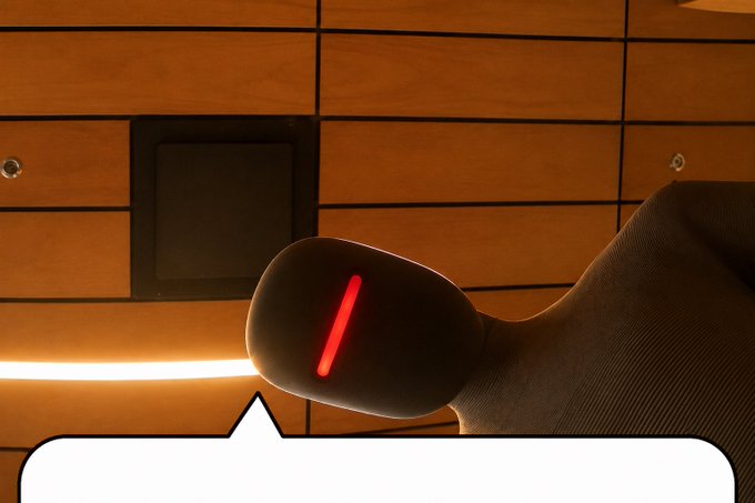 A close-up photo of a black robotic device with a red glowing slit, positioned in front of a wooden paneled wall with a partial speech bubble at the bottom.