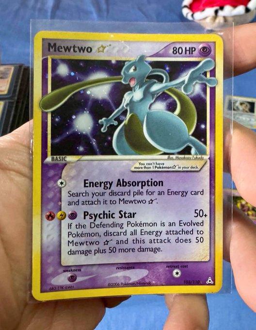 A hand holding a holo Mewtwo Pokémon trading card (80 HP) with attacks 'Energy Absorption' and 'Psychic Star 50+', stored in a protective sleeve.