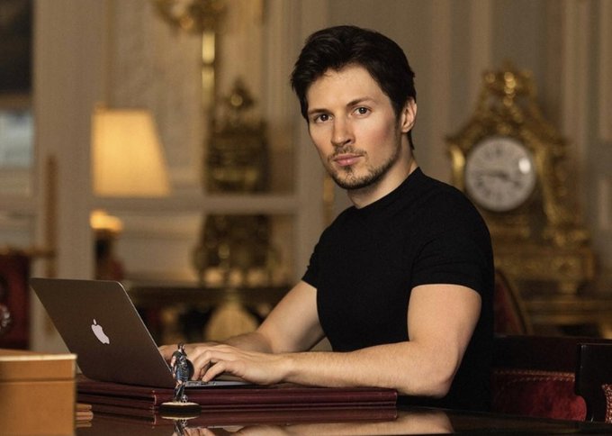 A young man with dark hair and a black t-shirt sits at an elegant desk using a silver Apple MacBook, with an ornate gold clock and warm lamp in the background.