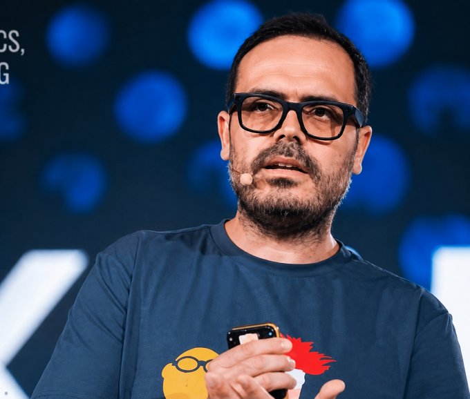 A man with glasses and a beard wearing a blue t-shirt speaking into a lapel microphone while holding a phone, on a stage with a dark blue bokeh background.