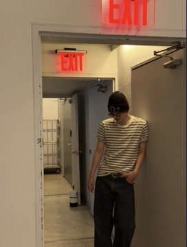 The image shows a young man wearing glasses, a striped shirt, and black pants standing in front of an exit door with his hand on his hip. The photo appears to have been taken by someone else or has been edited for use elsewhere. The caption provided suggests that this image might be related to the post #LEO #리오".
