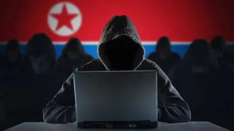 A hooded figure uses a laptop with their face obscured, set against a backdrop featuring the North Korean flag and shadowy silhouettes of other hooded individuals.