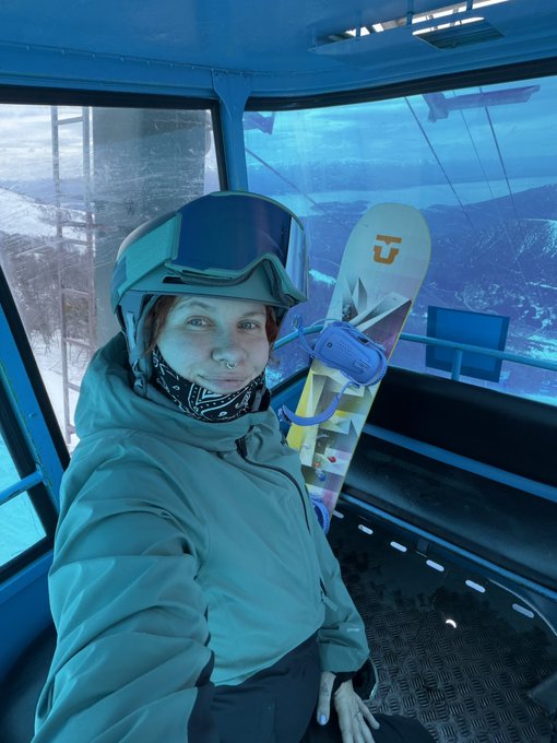 A selfie of a person wearing a grey winter jacket and ski goggles perched on their helmet, seated inside a blue ski lift gondola with snowboards visible in the background.