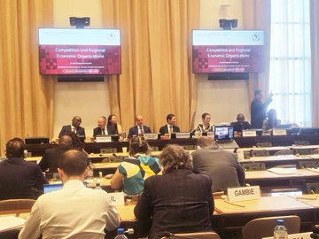 IOTA representatives participate in a panel at the Competitive Regional Economic Policies seminar in Geneva, as indicated by the screens behind the speakers. The "GAMBÉ" placard on a participant's desk suggests an African delegation, highlighting IOTA's engagement in international economic policy discussions.