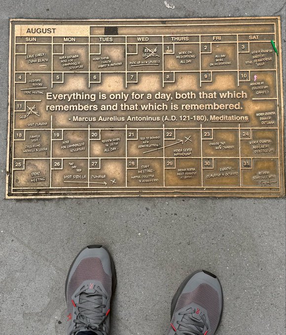 A worn August calendar lies on the ground with handwritten notes, overlaid with the quote 'Everything is only for a day, both that which remembers and that which is remembered' by Marcus Aurelius Antoninus, with a pair of gray sneakers visible at the bottom.