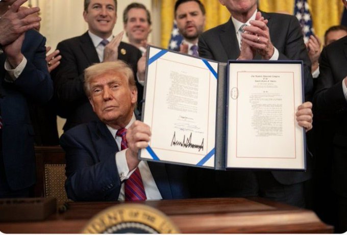Donald Trump holds up a signed executive order in the White House, displaying documents with "Donald J. Trump" visible, surrounded by applauding officials. The image illustrates the post's point about executive actions, showing a successful last-minute signing ceremony that the caption compares to the precarious legislative journey of the Clarity Act and Genius Act.