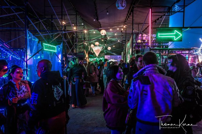 The image shows a large group of people gathered together, possibly at an event or festival with neon lights illuminating the area. The crowd appears to be engaged in conversation and enjoying themselves. There are several individuals standing close to each other, while others are spread out throughout the scene. A few people can also be seen holding backpacks, indicating that they might have brought personal belongings with them for the occasion. The overall atmosphere seems lively and social, as everyone is participating in or observing the event together.