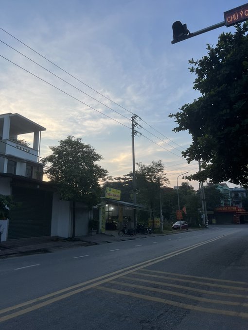 A street scene at dusk with a partly cloudy sky, showing a road with yellow markings, power lines, a traffic camera, trees, and buildings including a sign partially reading "CHỢ C" in the upper right.