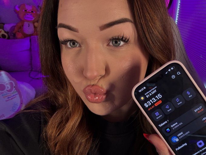 A woman with makeup making a kissing face while holding up a phone showing a crypto trading app with $313.15 and tokens including Solana and USDC, against a purple-lit background with stuffed animals.