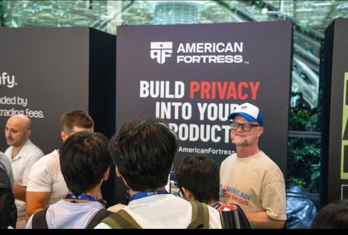 A trade show booth for 'AMERICAN FORTRESS' displays the slogan 'BUILD PRIVACY INTO YOUR PRODUCT' as attendees converse with a representative wearing a branded cap.