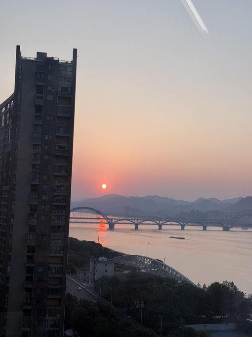 A sunset scene featuring a tall apartment building on the left, a bridge spanning a river, and silhouetted mountains with the orange sun on the horizon.
