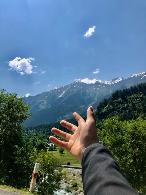 A first-person view of a hand reaching out toward a stunning mountain landscape with snow-capped peaks, lush green trees, and a river valley below under a bright blue sky.