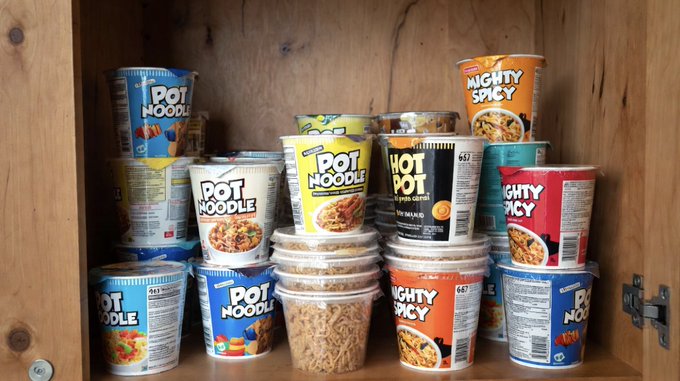 A cupboard shelf stocked with various Pot Noodle and Mighty Spicy instant noodle cups in colorful packaging.