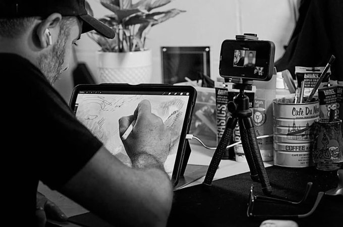 A black and white photo of a man wearing a cap and earbuds drawing on a tablet with a stylus, while a smartphone on a tripod records his artistic process.