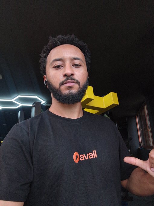 A bearded man wearing a black 'Avail' t-shirt poses for a selfie in what appears to be a gym, pointing at the Avail logo on his shirt.