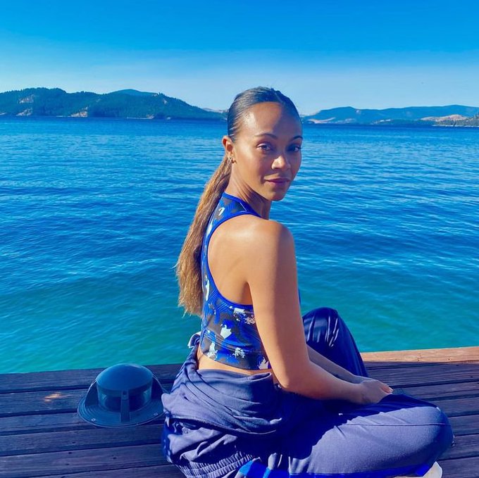 A woman with a long ponytail sits on a wooden dock beside a bright blue lake, wearing a blue patterned top with a dark blue garment draped around her legs.