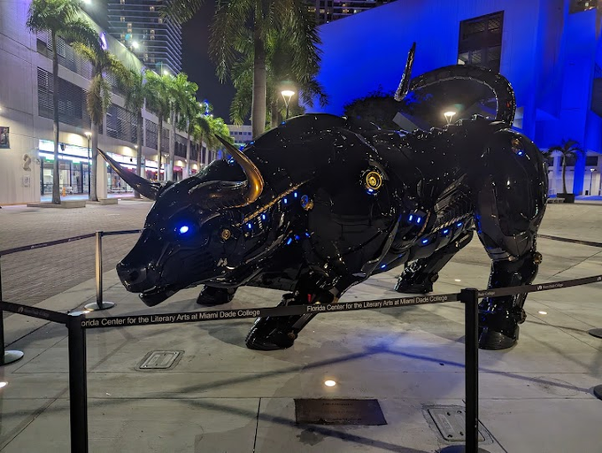 A large metallic bull sculpture with glowing blue eyes stands on a city sidewalk at night, displayed behind a rope barrier with signage for the 'Florida Center for the Literary Arts at Miami Dade College'.