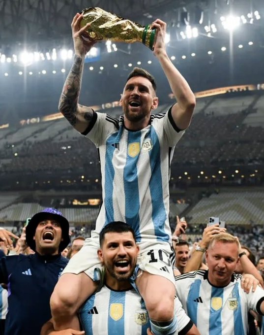 Lionel Messi in Argentina's jersey holding up the FIFA World Cup trophy while being carried on a teammate's shoulders in a stadium celebration.