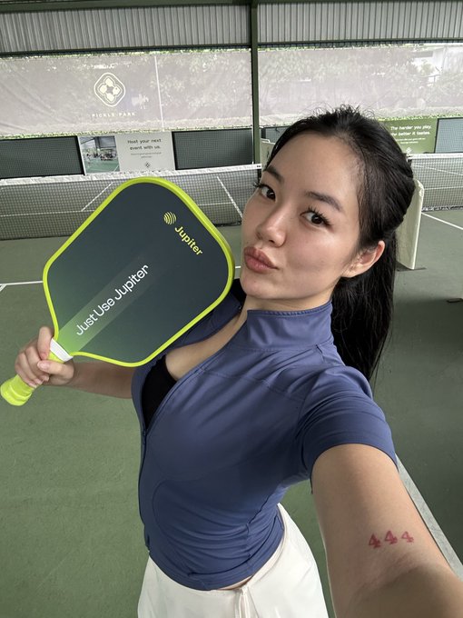 A woman taking a selfie in a pickleball court while holding a green and black paddle that says 'Just Use Jupiter'.