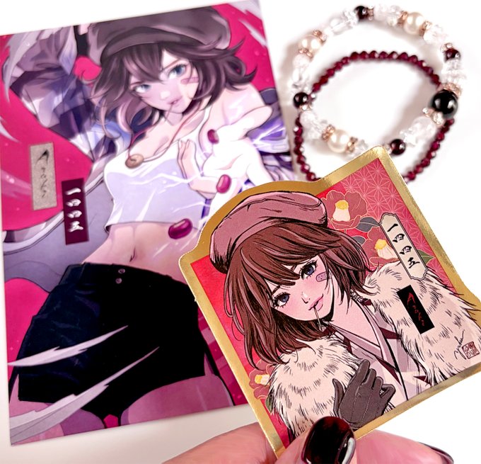 The image features a hand holding up two items, one being a sticker with an illustration of a girl wearing a hat and another item being a bracelet. The background appears to be pink, which complements the overall color scheme of the objects held by the person. The caption on the image reads "GM! My birthstone is red And my Azuki background is red too A coincidence, or maybe destiny I’ll keep this color close like a lucky charm and move forward in my own way". This suggests that the hand holding up these items might be expressing their connection to their birthstones or Azukas.