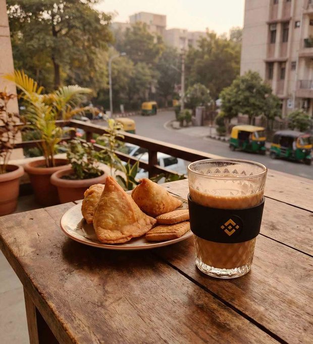 A glass of chai beside a plate of samosas on a wooden balcony table, with auto-rickshaws visible on the street below in what appears to be an Indian neighborhood. The image captures the classic monsoon-evening pairing the post references, while subtly placing the items next to a Binance-branded sleeve—a visual nod to the question about trading's most iconic combo.

**Visible text/logo:** "BINANCE" lettering and logo on the black sleeve around the glass.